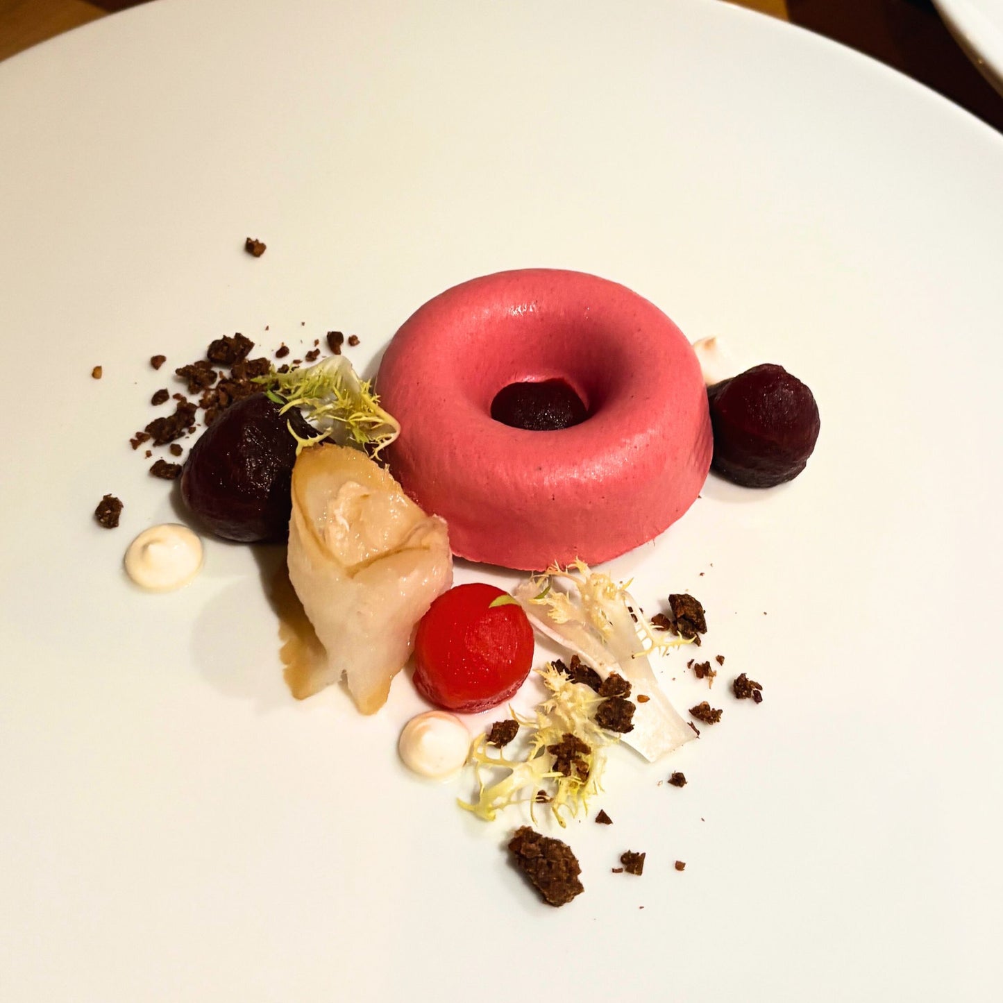 Fein angerichtetes Gourmet-Dessert mit rosa Mousse in Ringform, begleitet von Roter Bete, Sauce, Crumbles und essbarer Dekoration auf weißem Teller.