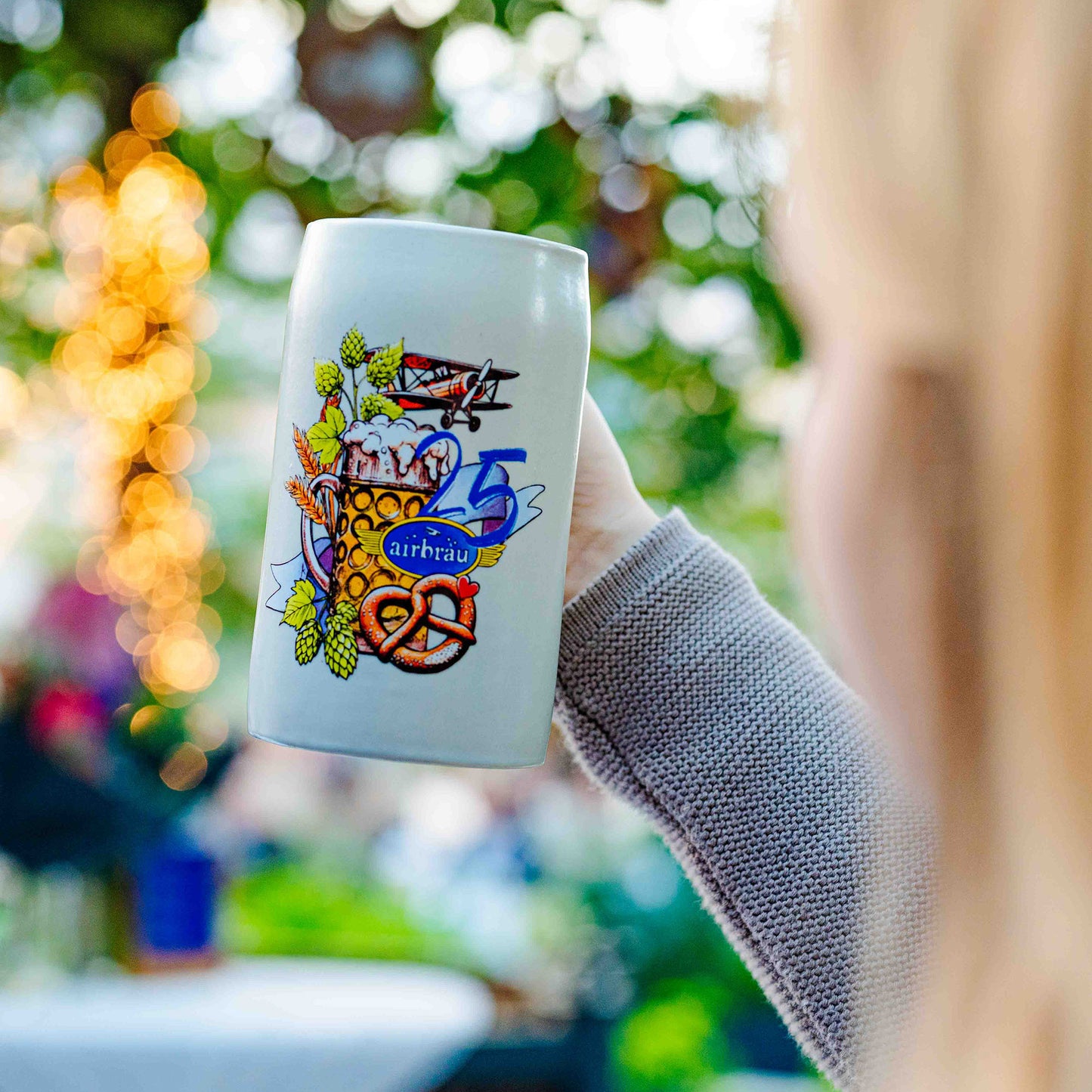 Airbräu-Krug mit farbigem Jubiläumsdesign „25 Jahre Airbräu“, Hopfen, Breze und Flugzeugmotiv . Zeigt Krug Hoch gehoben.