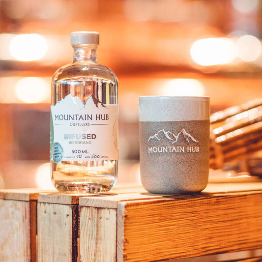 Flasche Mountain Hub Distillers Infused Bierbrand (500 ml) mit klarem Inhalt neben einem grauen Keramikbecher mit Mountain Hub Logo, arrangiert auf rustikalem Holzuntergrund vor warmem, unscharfem Hintergrund.