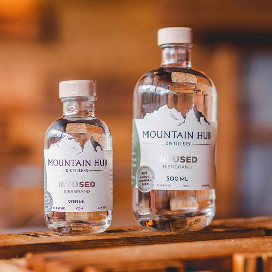 Zwei Flaschen Mountain Hub Distillers Infused Bierbrand (200 ml und 500 ml) mit klarem Inhalt und weißem Etikett, präsentiert auf rustikalem Holzuntergrund.
