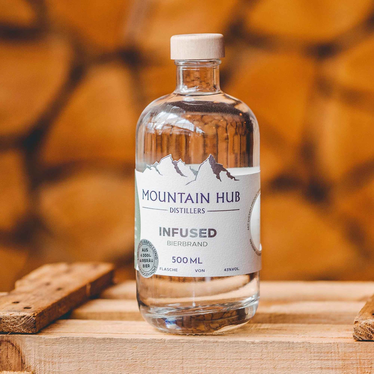 Flasche Mountain Hub Distillers Infused Bierbrand (500 ml) mit schlichtem weißen Etikett und Holzverschluss, präsentiert auf Holzuntergrund vor gestapeltem Holz im Hintergrund.