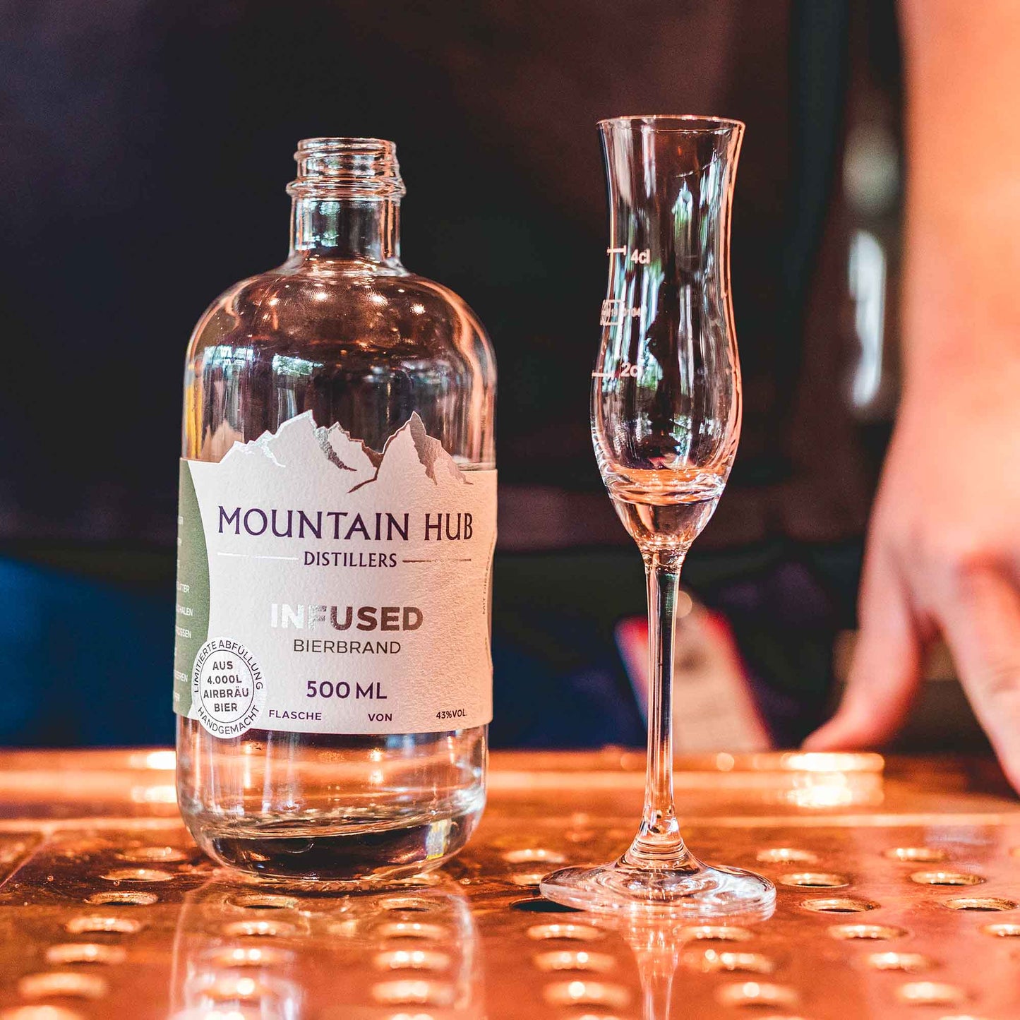 Flasche Mountain Hub Distillers Infused Bierbrand (500 ml) neben einem schmalen Schnapsglas auf einer glänzenden, kupferfarbenen Oberfläche, im Hintergrund unscharfe Person.