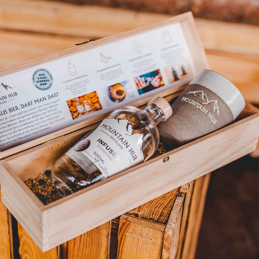 Geschenkbox aus Holz mit einer Flasche Mountain Hub Distillers Infused Bierbrand (500 ml) und einem grauen Keramikbecher mit Mountain Hub Logo, auf rustikalem Holzuntergrund.