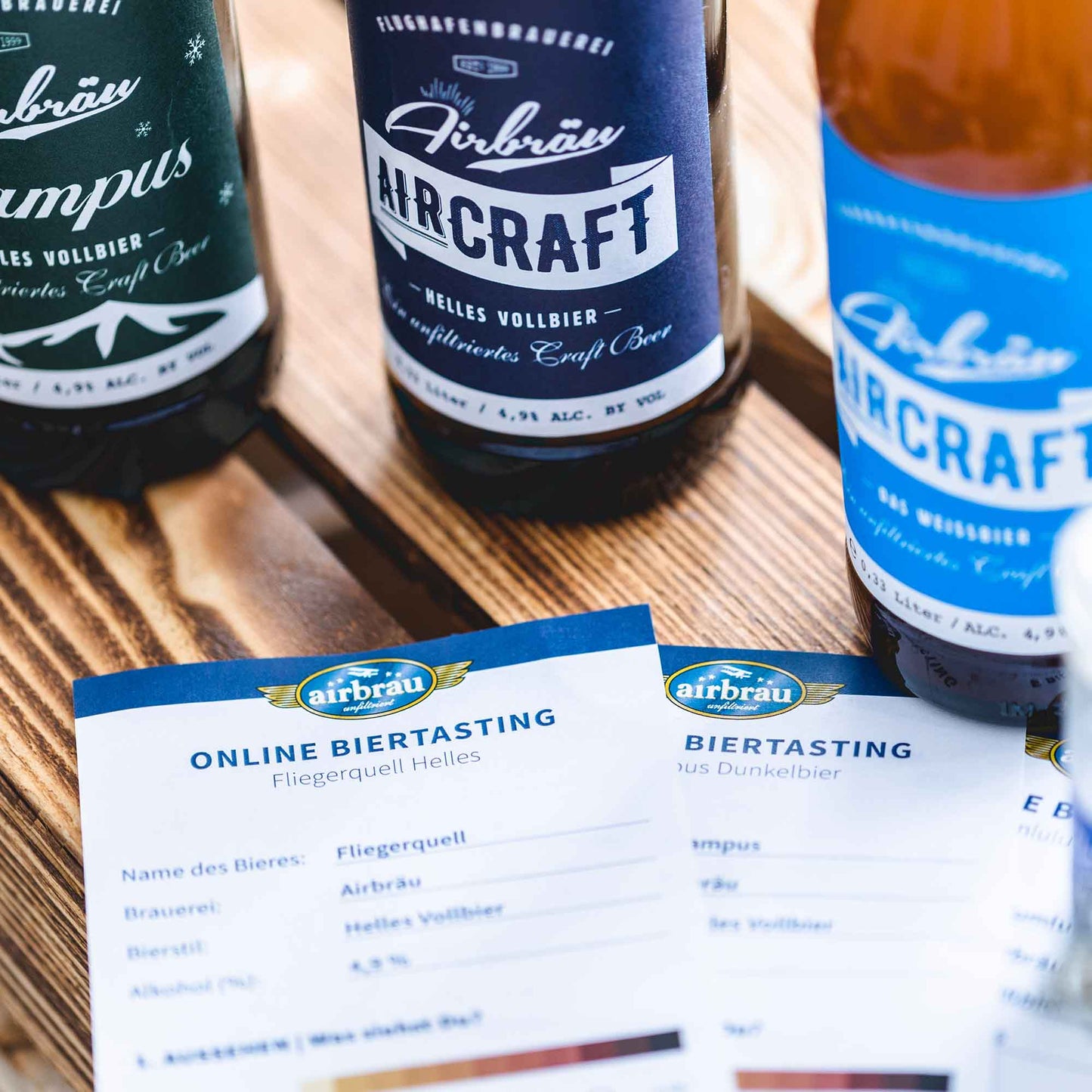 Verschiedene Airbräu-Bierflaschen, darunter „Aircraft“ und „Campus“, zusammen mit Informationskarten für ein Online-Biertasting auf einem Holztisch.
