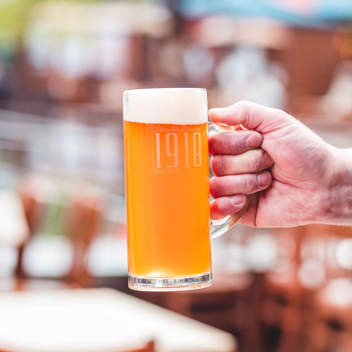 Hand hält einen gefüllten Bierkrug mit dem Schriftzug „1918“ und Schaumkrone, unscharfer Biergarten-Hintergrund mit warmen Farbtönen.