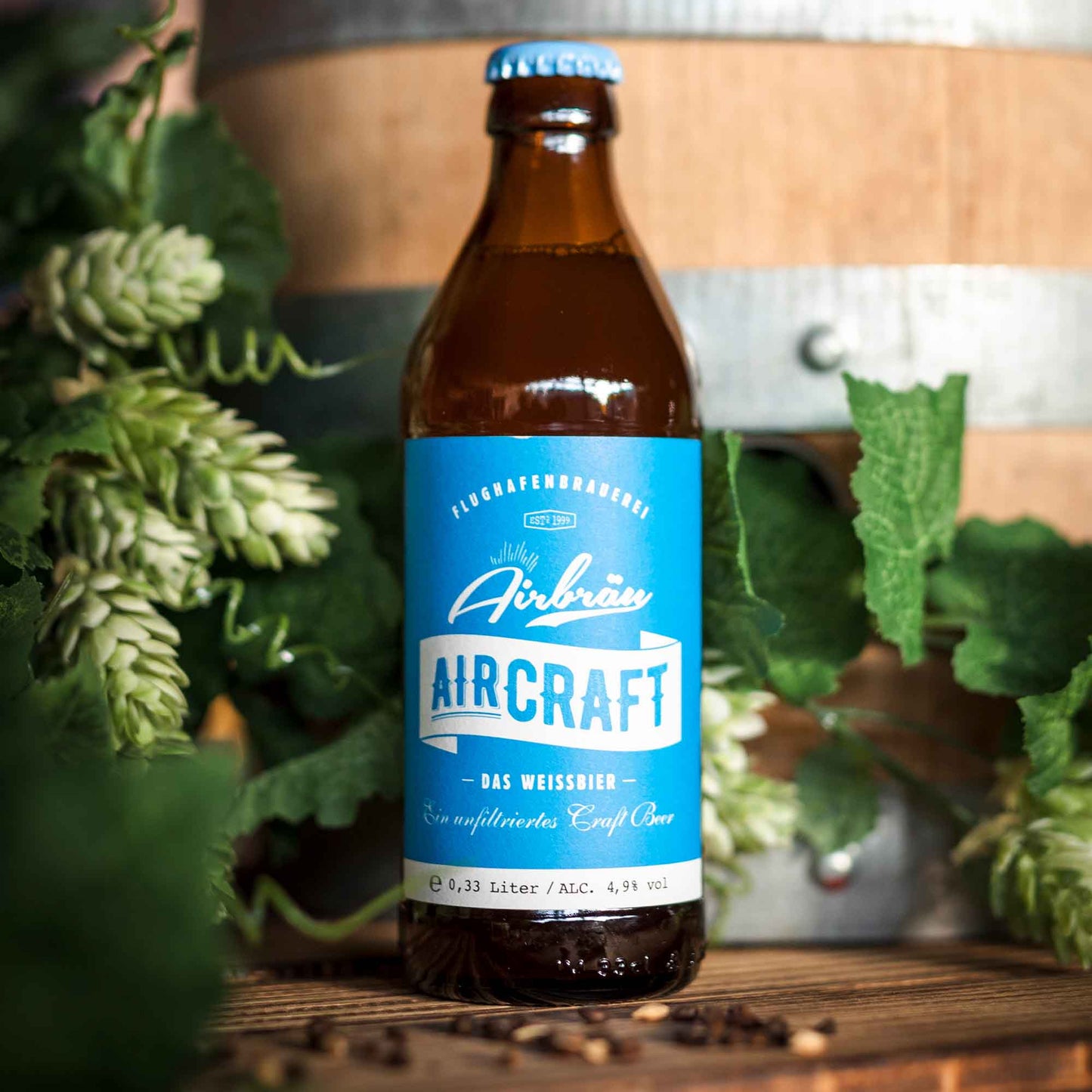 Flasche Airbräu Aircraft Das Weissbier mit blauem Etikett vor einem Holzfass, umgeben von frischen Hopfendolden und grünen Blättern auf einem Holztisch.