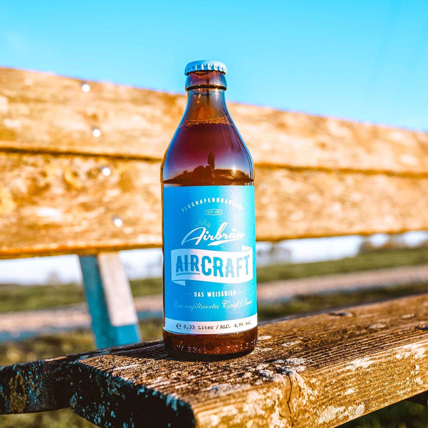 Flasche Airbräu Aircraft Das Weissbier auf einer verwitterten Holzbank im Freien, vor blauem Himmel und grüner Landschaft.