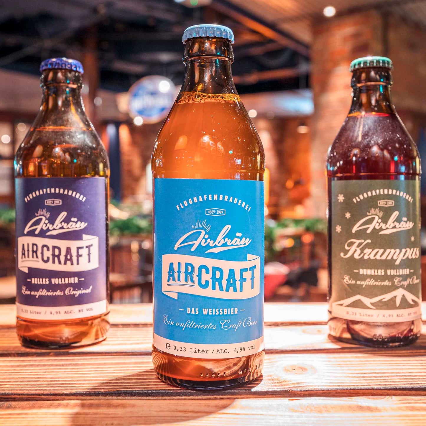 Drei Airbräu-Bierflaschen auf einem Holztisch in gemütlicher Brauhaus-Atmosphäre, darunter „Aircraft Helles Vollbier“, „Aircraft Das Weissbier“ und „Krampus Dunkles Vollbier"