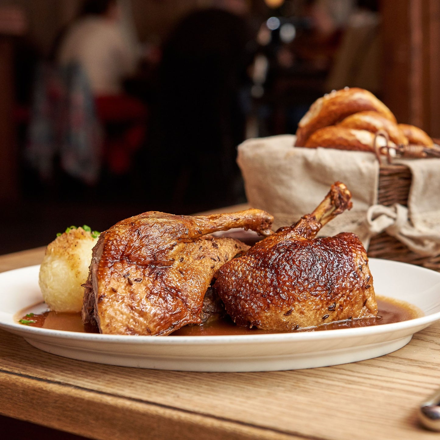 Ein Teller mit knusprig gebratener Gänsekeule in dunkler Soße, serviert mit einem Kartoffelknödel. Im Hintergrund steht ein Brotkorb mit frischen Brezen auf einem Holztisch in gemütlicher Wirtshaus-Atmosphäre.