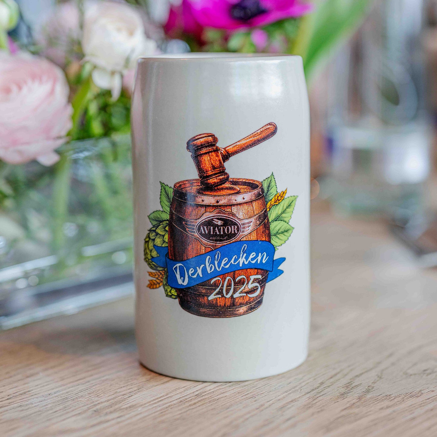 Ein heller Steinzeug-Bierkrug steht auf einem Holztisch. Auf der Vorderseite ist eine farbige Illustration eines Holzfasses mit Richterhammer, Hopfenblättern und Ähren sowie der Aufschrift „Aviator – Derblecken 2025“ auf einem blauen Banner zu sehen. Im Hintergrund befinden sich unscharf arrangierte Blumen in Vasen.