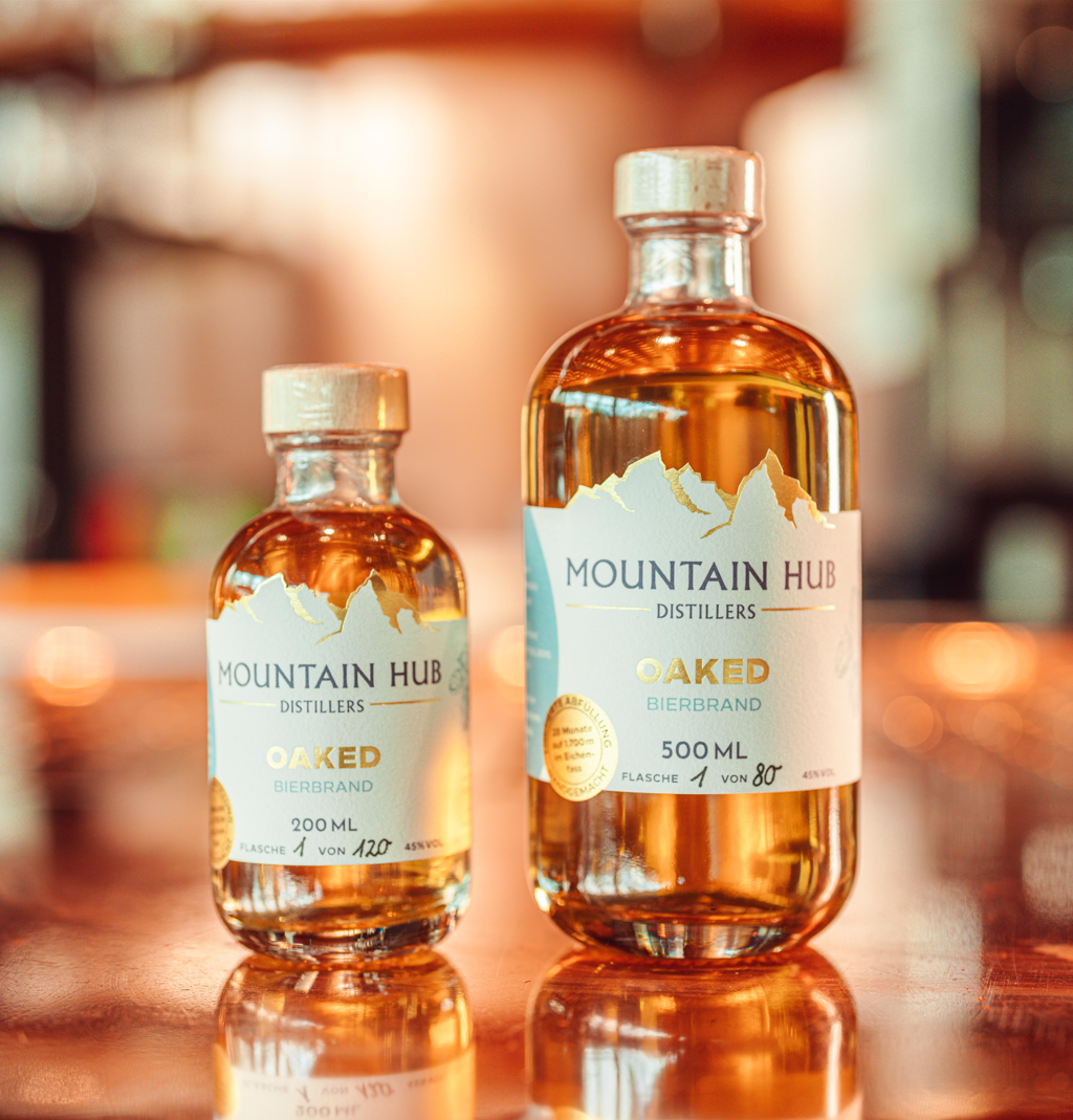 Zwei Flaschen Mountain Hub Distillers Oaked Bierbrand (200 ml und 500 ml) mit goldbraunem Inhalt und weiß-goldenem Etikett, auf glänzendem Untergrund vor unscharfem Hintergrund.