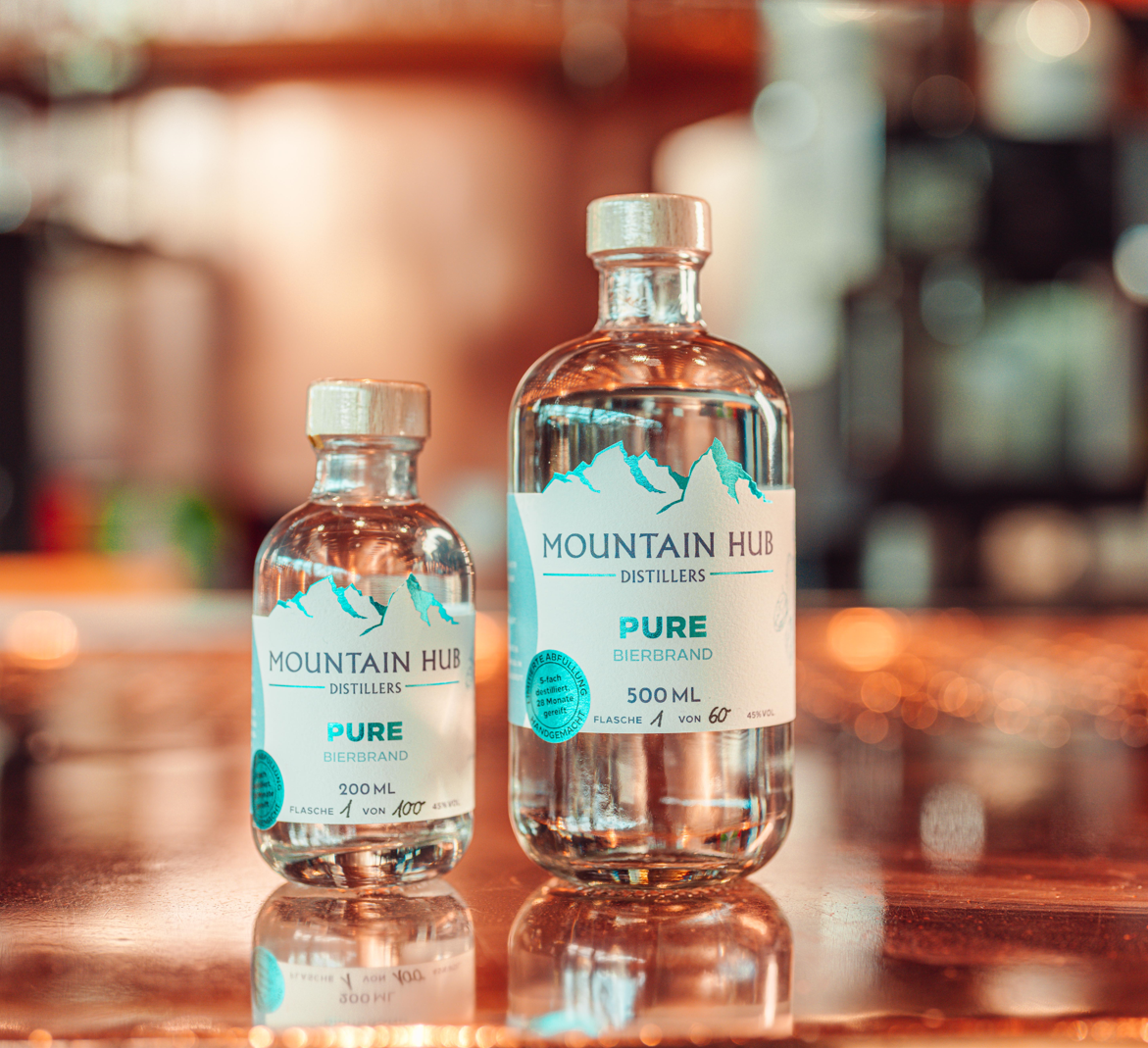 Zwei Flaschen Mountain Hub Distillers Pure Bierbrand (200 ml und 500 ml) mit klarem Inhalt und weiß-türkisfarbenem Etikett, auf glänzendem Untergrund vor unscharfem Hintergrund.