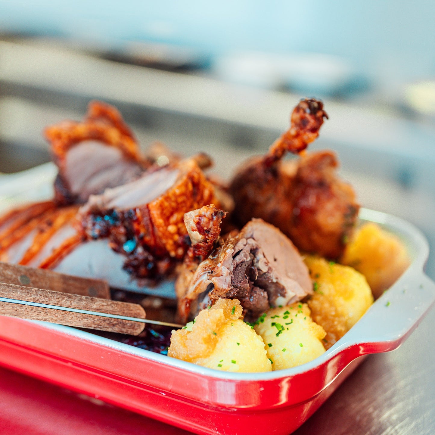 Herzhaft angerichteter Braten mit knuspriger Kruste und saftiger Fleischfüllung, serviert mit Kartoffelknödeln in einer roten Auflaufform, auf einem Edelstahltisch.