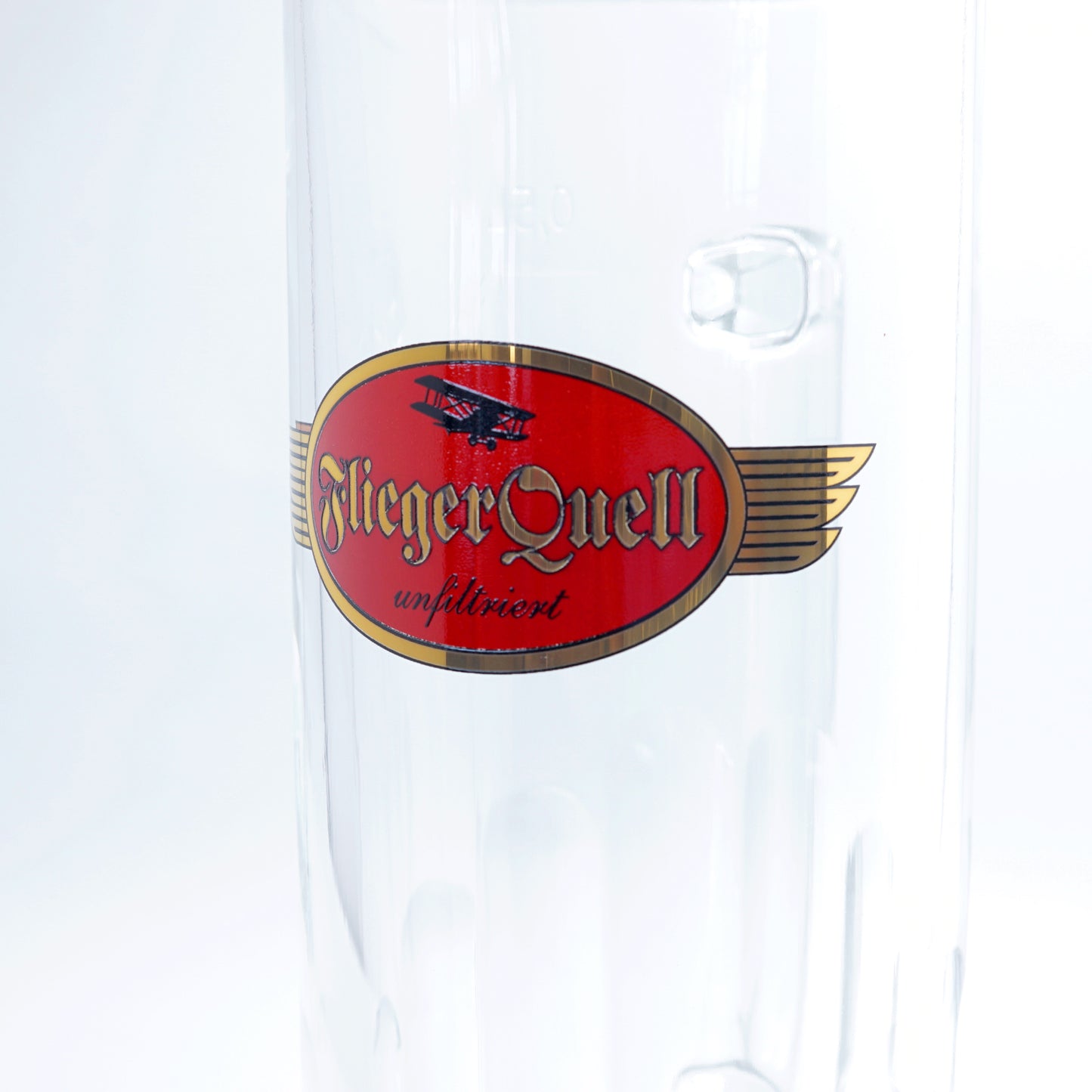 Nahaufnahme eines 0,5-Liter-Bierkrugs mit rotem und goldenen FliegerQuell-Logo und der Aufschrift „unfiltriert“, auf weißem Hintergrund. Leicht nach Links gedreht.