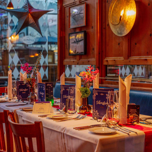 Festlich gedeckter Tisch mit Menükarten für Silvester 2025, weißen Stoffservietten, Blumen und gläserner Tischdekoration.