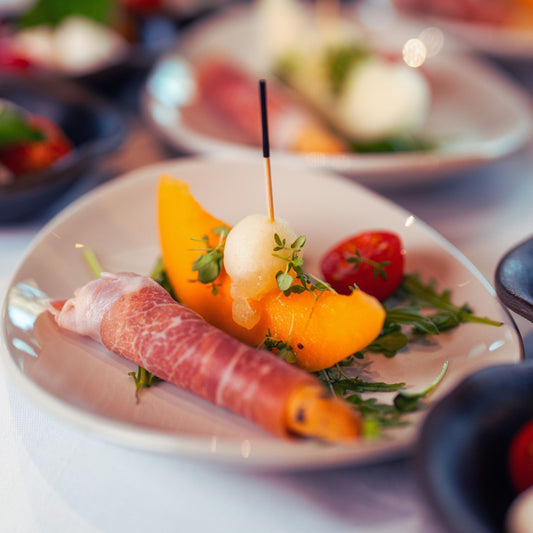 Vorspeisenteller mit Schinkenröllchen, Melonenspalte und einer kleinen Kugel Mozzarella auf Rucola.
