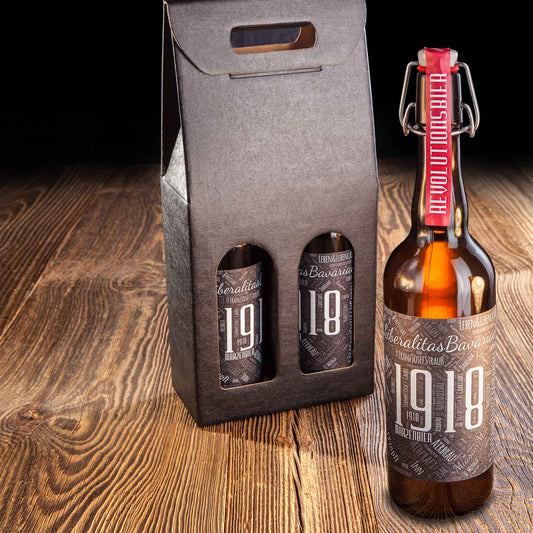 Flasche Airbräu 1918 Märzenbier mit Bügelverschluss und rotem Band „Revolutionsbier“ neben schwarzem Tragekarton mit zwei weiteren Flaschen, auf dunkler Holzoberfläche.