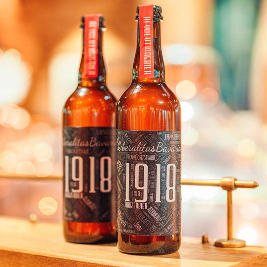 Zwei Flaschen Airbräu 1918 Märzenbier mit schwarzem, grafisch gestaltetem Etikett und rotem Halsband mit der Aufschrift „Revolutionsbier“, vor unscharfem, warmem Hintergrund.