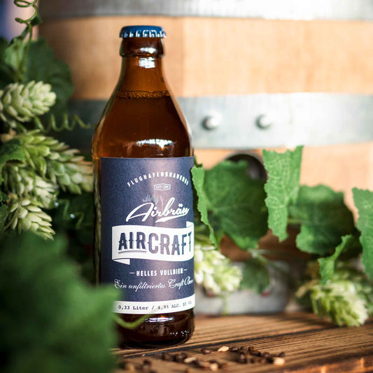 Flasche Airbräu Aircraft Helles Vollbier vor einem Holzfass, umgeben von frischen Hopfendolden und grünen Blättern auf einem Holztisch.
