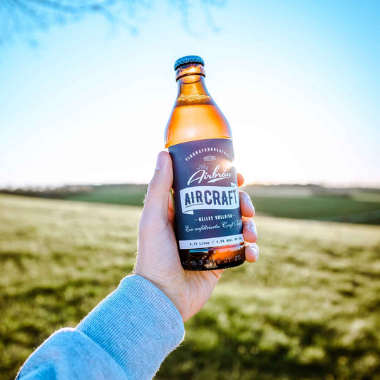Hand hält eine Flasche Airbräu Aircraft Helles Vollbier vor einer sonnigen, grünen Landschaft mit klarem Himmel im Hintergrund.