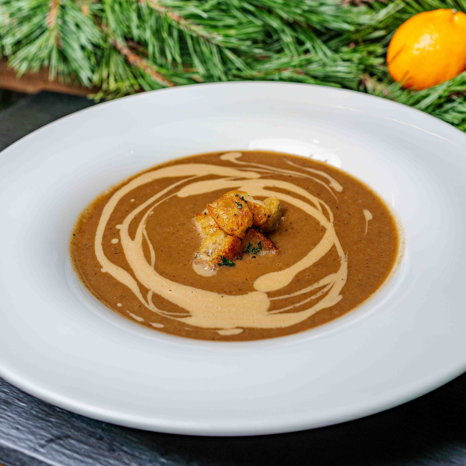 Cremige Maronensuppe mit feinem Sahnespiegel und knusprigen Croûtons, serviert in einem weißen tiefen Teller – winterlich angerichtet mit Tannenzweigen im Hintergrund.