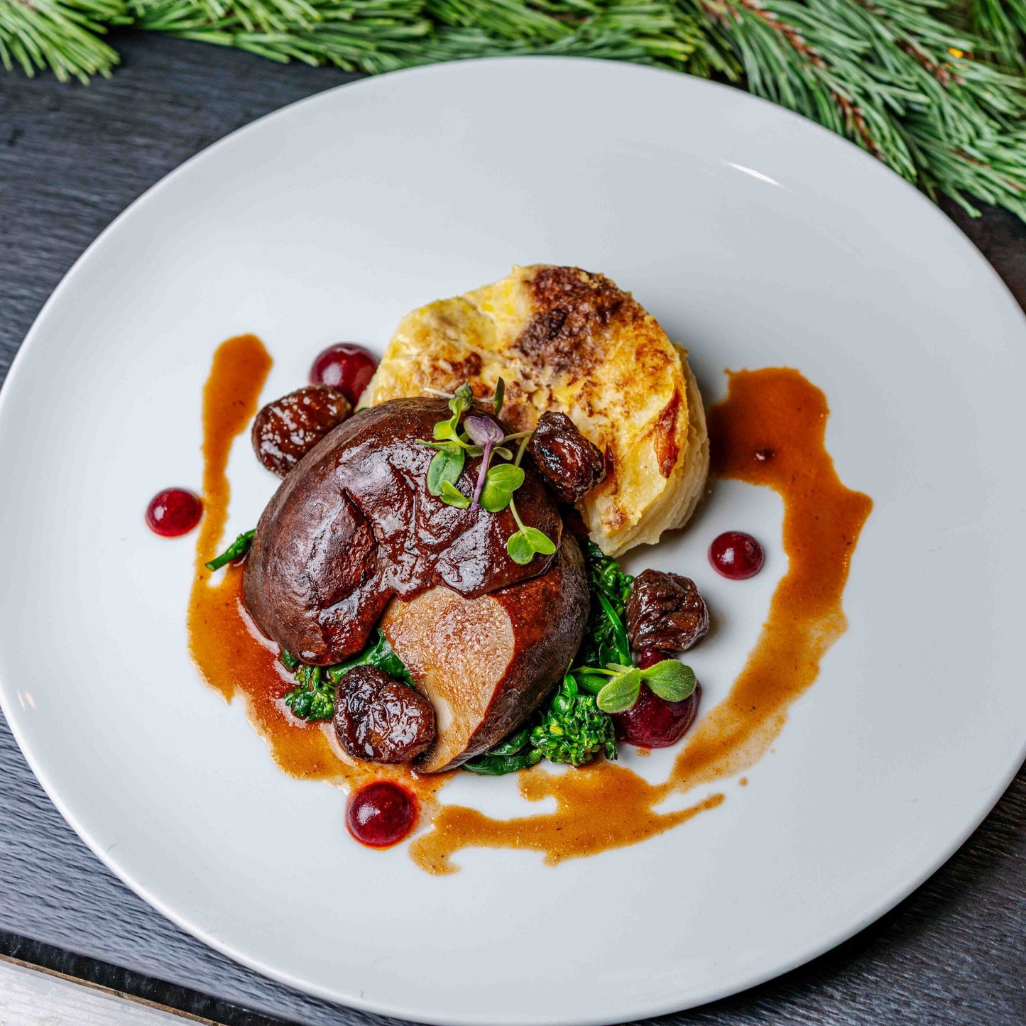 Veganes Portobello-Steak mit Thymianjus, wildem Brokkoli, cremigem Kartoffelgratin und glasierten Maronen, kunstvoll angerichtet auf einem weißen Teller.