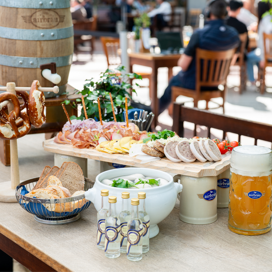Stammtisch-Package im Airbräu: rustikales Brotzeitbrettl mit Wurst- und Käsespezialitäten, Brezen, Weißwürste im Terrinentopf, Brotkorb, kleine Enzianschnäpse und eine frisch gezapfte Maß Bier auf einem Tisch im Biergarten.
