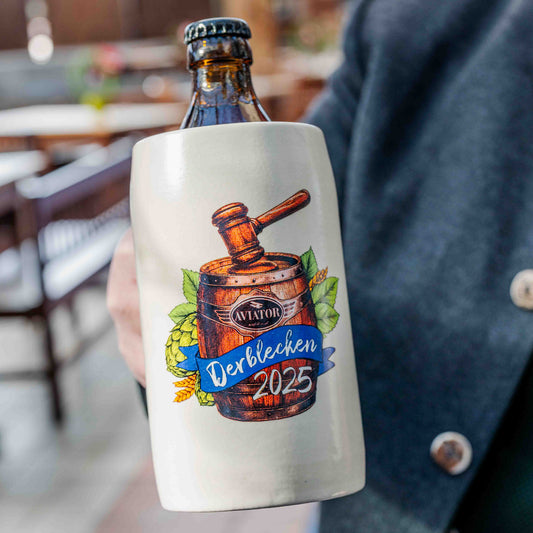 Ein traditioneller Bierkrug aus hellem Steinzeug wird in der Hand gehalten. In ihm steckt eine braune Glasflasche mit Kronkorken. Auf dem Krug ist ein farbiges Motiv mit einem Holzfass, einem Richterhammer, Hopfenblättern und der Aufschrift „Aviator – Derblecken 2025“ abgebildet.