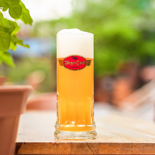 Gefülltes 0,5-Liter-Bierglas mit rotem und goldenen FliegerQuell-Logo sowie cremigem Bierschaum, auf einem Holztisch mit grünem, unscharfem Hintergrund.