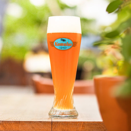 Gefülltes 0,5-Liter-Weißbierglas mit Kumulus-Logo, gold-orangefarbenem Bier und cremigem Schaum, auf einem Holztisch mit grünem, unscharfem Hintergrund.
