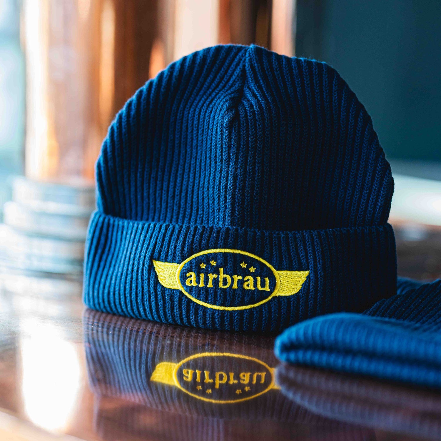 Dunkelblaue Strickmütze mit gelbem „airbräu“-Logo und gestickten Sternen, präsentiert auf einer glänzenden Holzoberfläche.