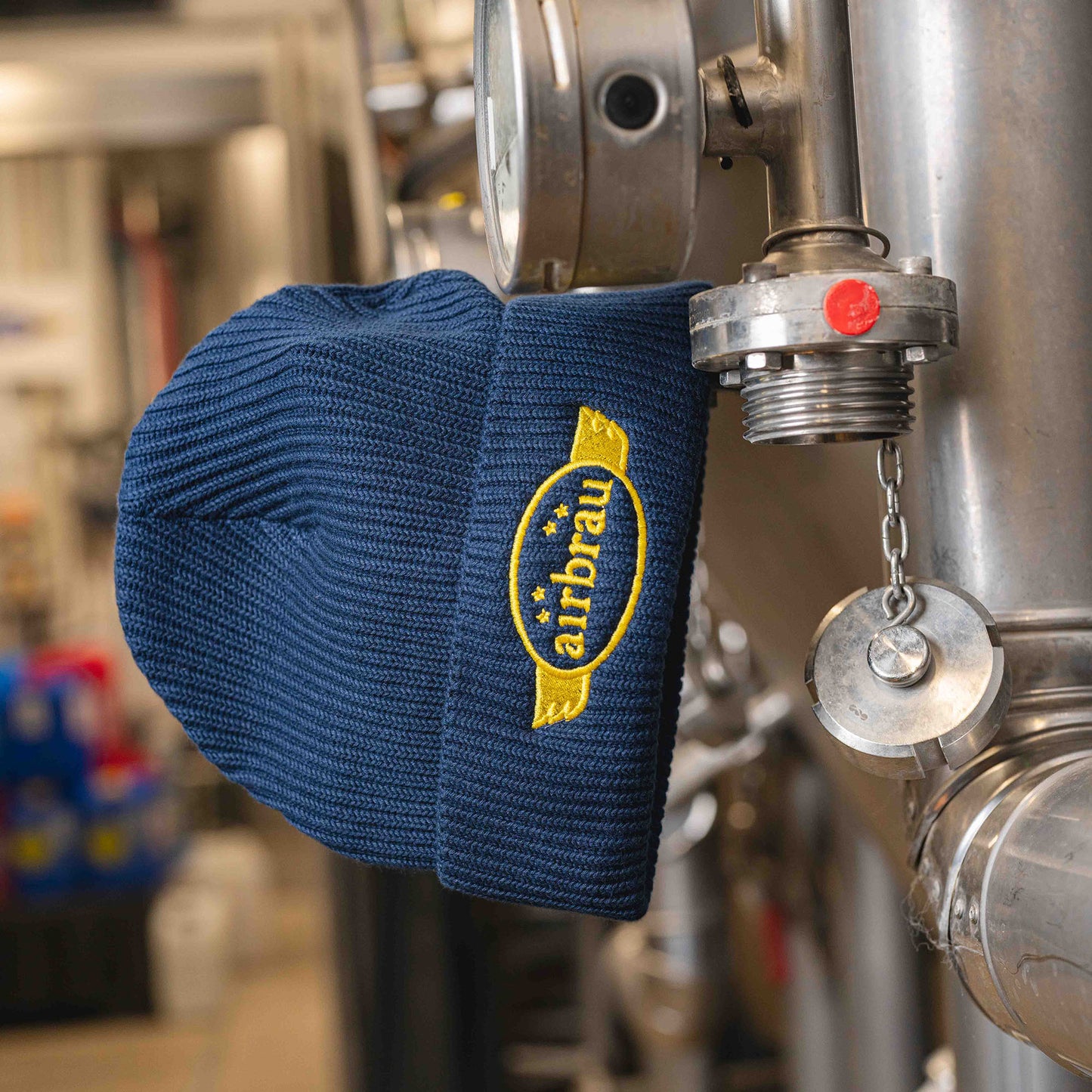 Dunkelblaue Strickmütze mit gelbem „airbräu“-Logo, aufgehängt an einem Edelstahl-Bierkessel in einer Brauerei.