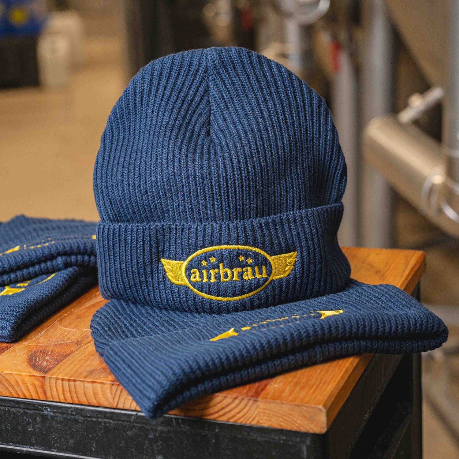 Dunkelblaue Strickmütze mit gelbem gesticktem Airbräu-Logo, präsentiert auf Holztisch mit weiteren Mützen im Hintergrund.