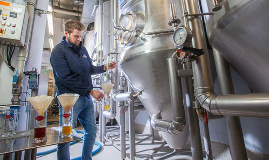 Brauer in der Brauerei zapft Proben aus einem großen Edelstahltank zur Qualitätskontrolle; mehrere Bierproben stehen in Gläsern mit Filtern auf einem Tisch bereit.